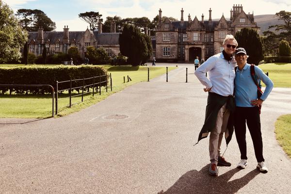 Two men are smiling and posing for a photo on a paved path, with a grand historic manor house visible in the background